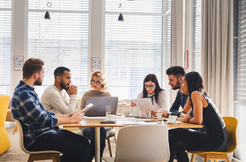 Six people in a bright office meeting around a table with papers, coffee, and laptop—focused and collaborative.