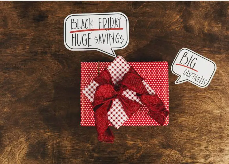 Polka-dot gift box with red bow on wooden surface, with speech bubbles reading 'Black Friday Huge Savings' and 'Big Discounts