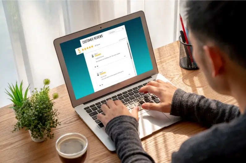 Person typing on laptop showing 5-star review; desk has plant, coffee, pens in sunlight.