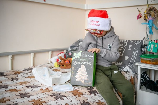 Boy in a Santa hat and festive glasses sits on a bed holding a gingerbread Christmas tree kit.